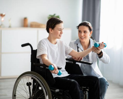 Physical rehabilitation concept. Young physiotherapist helping teenage boy in wheelchair to do exercises at home. Disabled adolescent training with dumbbells, working out his muscles indoors